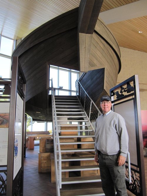 A man stands beside a staircase leading up to a large wooden boat structure inside a building.