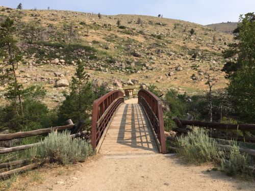 A wooden bridge spans a dirt path, surrounded by grassy hills and scattered trees in a natural landscape.