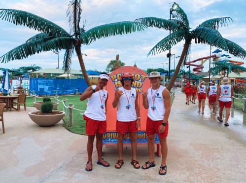 Three lifeguards in matching outfits pose in front of palm trees at a water park, smiling and making hand gestures.