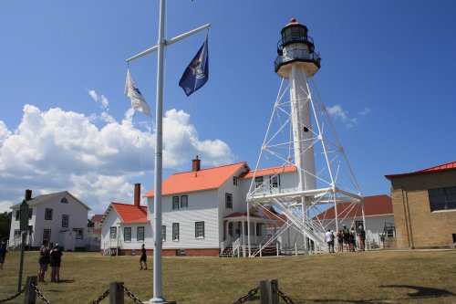 A white lighthouse with a black top stands beside historic buildings and flags under a blue sky with fluffy clouds.