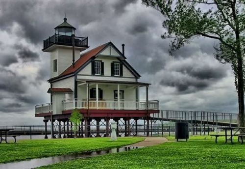 A charming lighthouse-style house on stilts, surrounded by green grass and a cloudy sky.