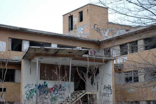 Abandoned building with graffiti on brick walls, overgrown vegetation, and a staircase leading to a boarded entrance.