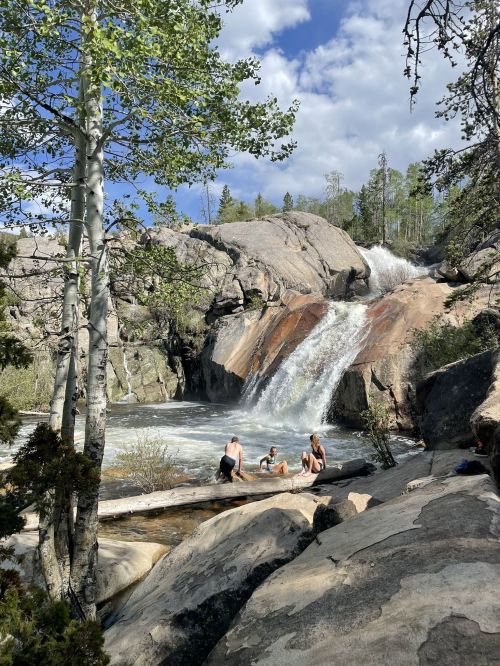 A scenic waterfall cascades over rocks, with people enjoying the water in a lush, green forest setting.