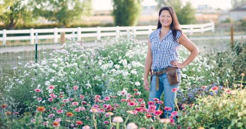 A smiling woman stands in a vibrant flower garden, wearing a checkered shirt and jeans, with a fence in the background.