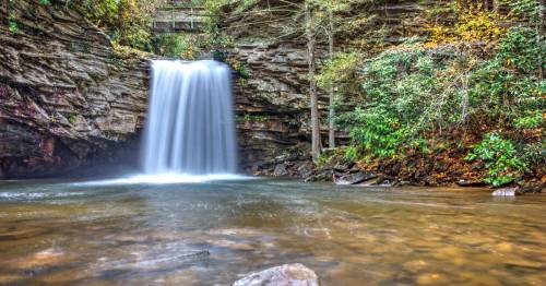 A serene waterfall cascades over rocky cliffs into a calm pool, surrounded by lush greenery and autumn foliage.