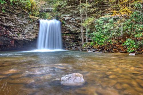 A serene waterfall cascades over rocky cliffs into a calm pool, surrounded by lush greenery and autumn foliage.