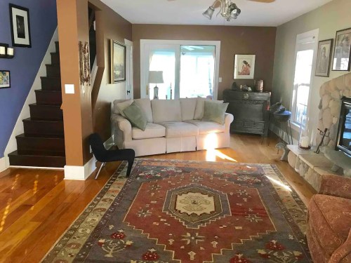 Cozy living room with a sofa, decorative rug, stone fireplace, and stairs leading to an upper level. Bright and inviting space.