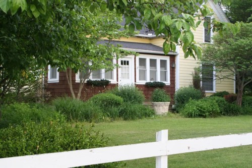 A charming house with a mix of yellow and brown siding, surrounded by lush greenery and a white picket fence.