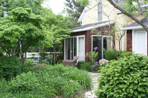 A cozy house surrounded by lush greenery and colorful flowers, featuring a screened porch and stone pathway.