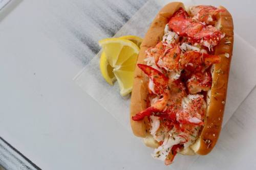A lobster roll filled with chunks of lobster meat, garnished with herbs, served with lemon wedges on the side.
