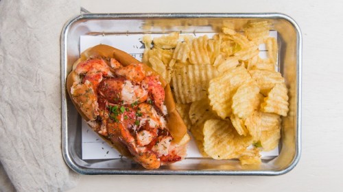A lobster roll on a bun with a side of ridged potato chips in a metal tray.