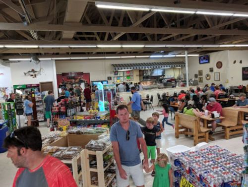 A busy indoor market with families, food counters, and various products displayed on shelves and tables.