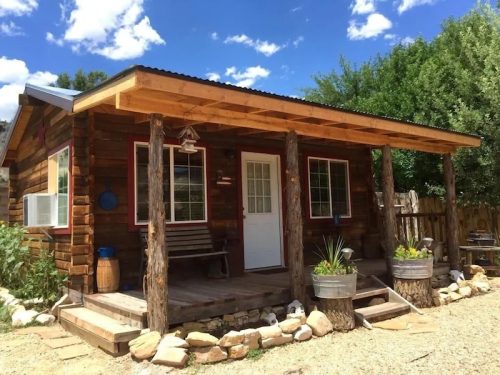 A cozy wooden cabin with a porch, surrounded by greenery and gravel, under a bright blue sky with fluffy clouds.
