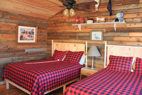 Cozy bedroom with two plaid beds, wooden walls, a ceiling fan, and rustic decor on shelves.