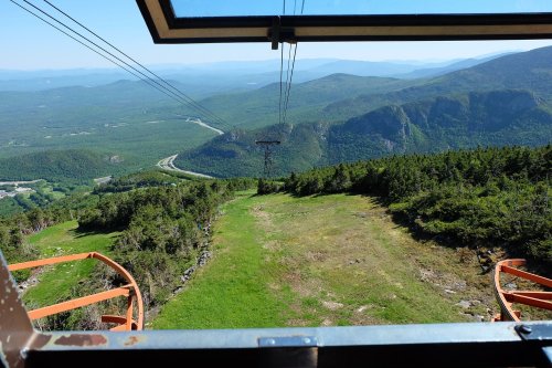 View from a cable car overlooking green mountains and a valley, with a clear blue sky in the background.