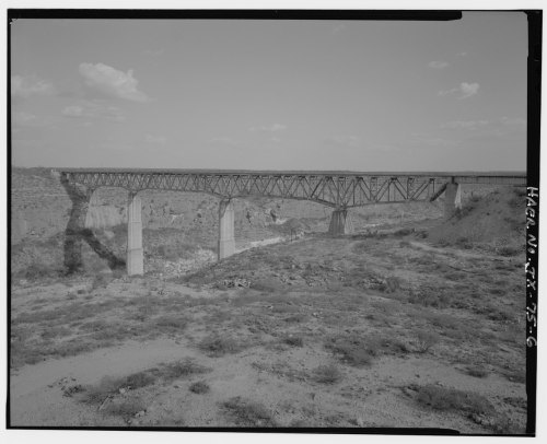 A large metal bridge spans a canyon, supported by tall concrete pillars, under a clear sky.