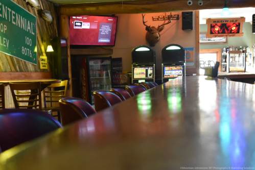 A rustic bar interior with wooden stools, a TV, and a deer head mounted on the wall, creating a cozy atmosphere.