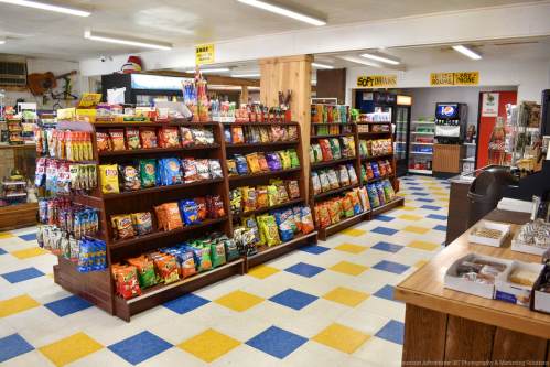 Interior of a convenience store featuring snack aisles, colorful tiled floor, and a beverage section in the background.