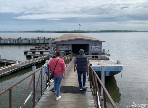 Two people walk along a wooden dock towards a restaurant on the water, with boats and a cloudy sky in the background.