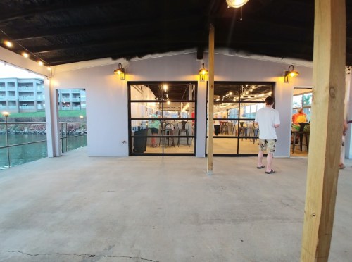 A waterfront building with large glass doors, outdoor seating, and warm lighting, overlooking a calm waterway.