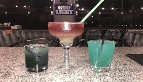 Three colorful cocktails on a bar counter: a large layered drink in a glass, and two smaller drinks in blue and green.