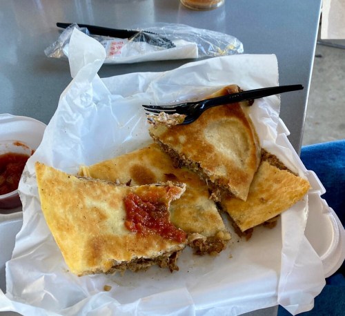 A partially eaten quesadilla with ground meat and salsa on a white paper plate, accompanied by a black fork.