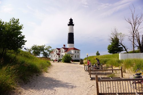 A sandy path leads to a black and white lighthouse surrounded by greenery and a historic building.