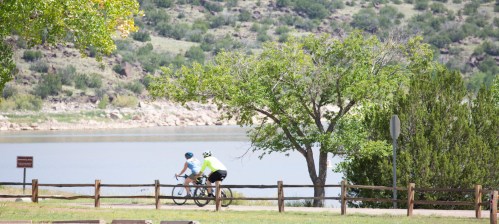 Two cyclists ride along a path near a lake, surrounded by greenery and hills in the background.