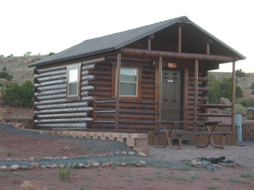 A small log cabin with a metal roof, surrounded by gravel and desert landscape, featuring a picnic table outside.