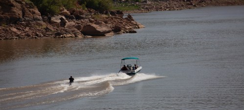 A boat pulls a person water skiing on a calm lake surrounded by rocky shores and greenery.