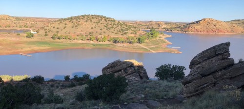 A serene landscape featuring a calm lake surrounded by hills and rocky formations under a clear blue sky.