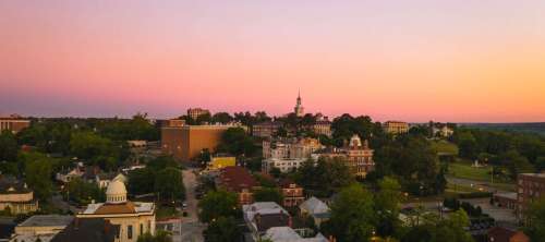 Aerial view of a town at sunset, featuring buildings, trees, and a colorful sky.