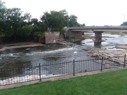 A river flows under a concrete bridge, with rocky banks and green grass in the foreground. Trees line the riverbank.