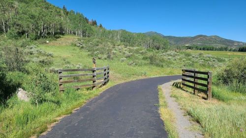 A paved path winds through a green landscape with wooden fences and mountains in the background under a clear blue sky.