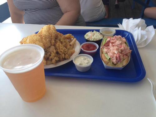 A tray with fried chicken, fries, coleslaw, a lobster roll, and a beer on a table.