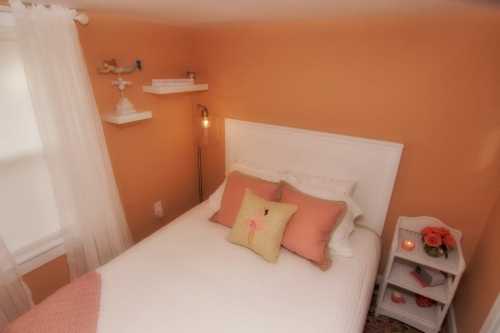 Cozy bedroom with an orange wall, white bed, decorative pillows, and a small nightstand with flowers.