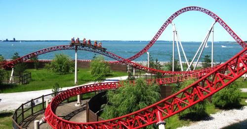A red roller coaster twists and turns over a scenic waterfront, with riders enjoying the thrill on a sunny day.