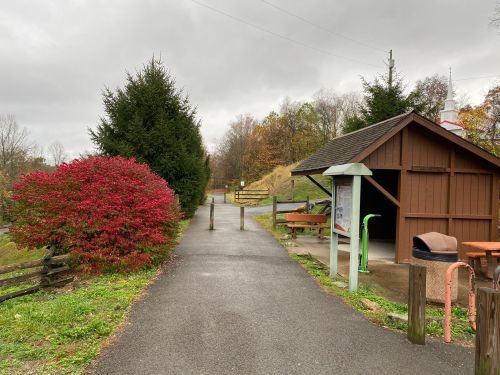 A paved path leads through a park with a red bush, a shelter, and trees under a cloudy sky.