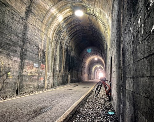 A bike leans against the wall of a dark tunnel, with light visible at the far end and textured stone walls.
