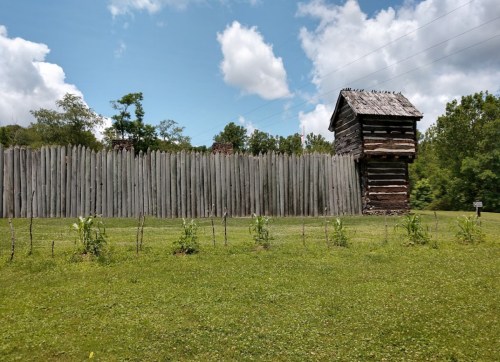 A wooden fort with a tall watchtower surrounded by a grassy area and trees under a partly cloudy sky.