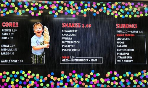A child holds a large ice cream cone, with a menu displaying cones, shakes, and sundaes prices and options.