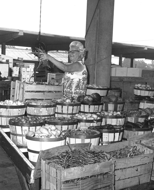 A woman with white hair holds up produce at a market, surrounded by baskets of fruits and vegetables.