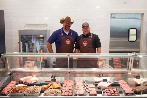 Two butchers in aprons stand behind a display case filled with various cuts of meat.