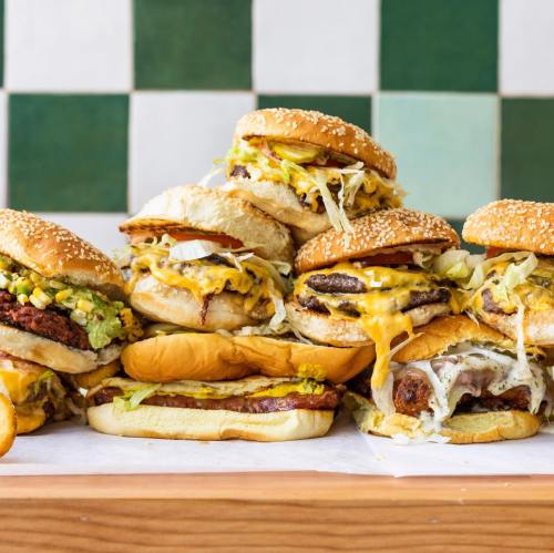 A stack of various burgers with toppings like cheese, lettuce, and sauces, set against a green tiled background.