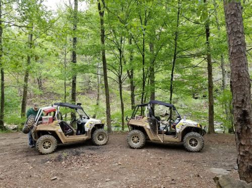 Two off-road vehicles parked on a dirt path surrounded by lush green trees and a creek in the background.