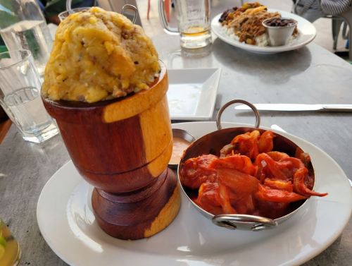 A wooden bowl filled with mashed plantains next to a small dish of spicy shrimp in a red sauce.