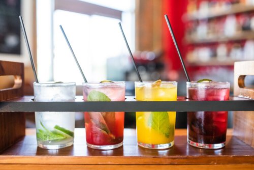 A wooden tray holds four colorful cocktails with straws, featuring mint and citrus garnishes, in a bright setting.