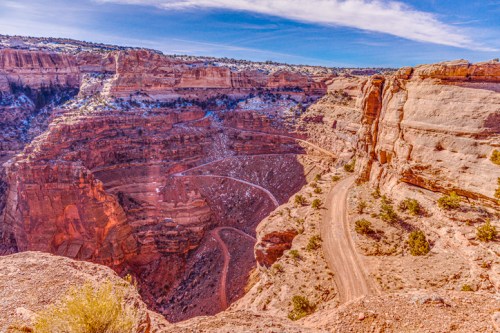 A scenic view of a winding dirt road through a deep canyon, surrounded by rocky cliffs and blue skies.