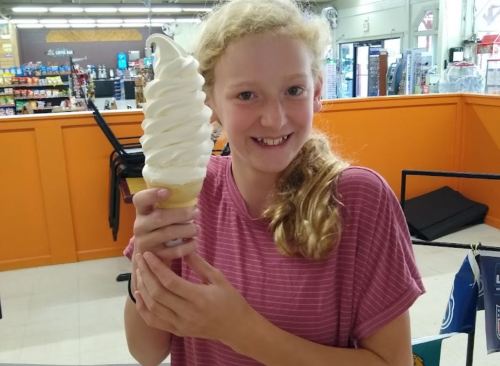 A smiling girl holds a large soft-serve ice cream cone in a colorful shop.