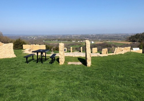A scenic view of a grassy area with stone structures and a picnic table, overlooking a valley under a clear blue sky.